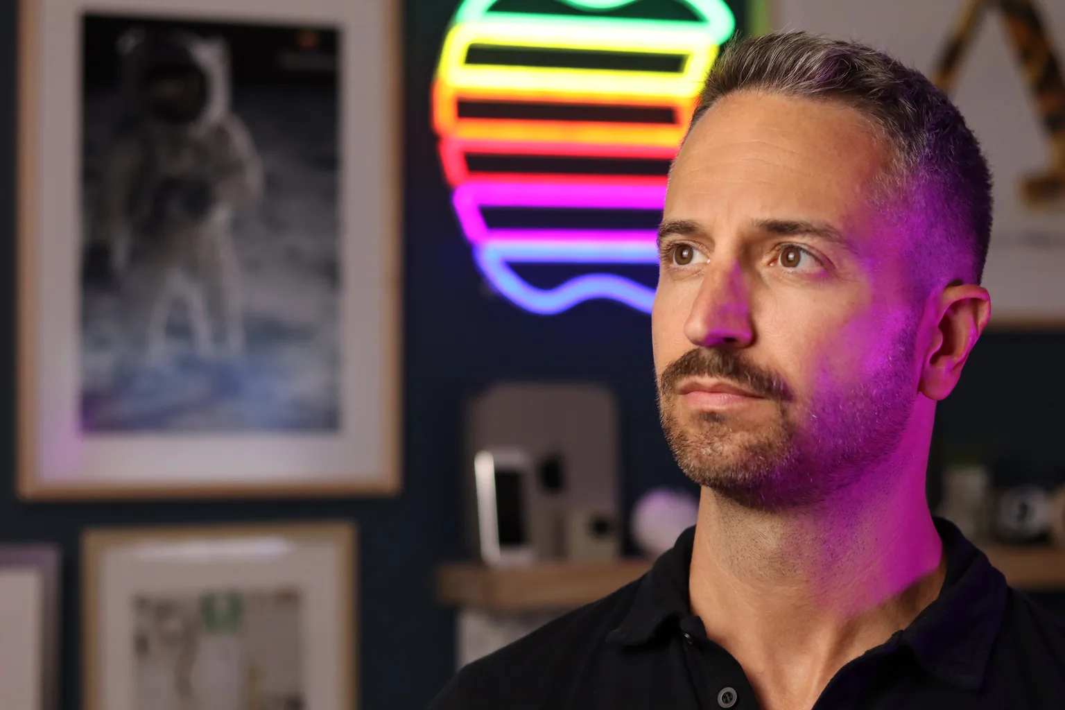 Christian Brescia in his studio, looking off-camera, lit in magenta by a large neon Apple logo on the wall behind him. An Apollo moonwalk photograph hangs to one side and a shelf of Apple devices sits below.
